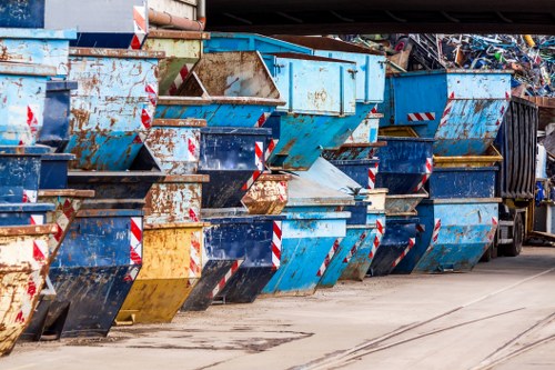 A row of large, industrial metal skip bins lined up outdoors on a paved surface, predominantly painted in shades of blue with some areas showing rust and wear. The bins are designed for rubbish collection and disposal, with open tops revealing accumulated debris inside. Some bins have red and white reflective warning stripes on the sides for safety visibility. The background includes occasional glimpses of other waste containers or equipment, indicating a waste management or disposal site, possibly in the vicinity of Chigwell or nearby areas. Weather conditions appear overcast, providing diffuse natural lighting across the scene, emphasizing the textures and colors of the metal skips. This image exemplifies typical rubbish removal operations managed by companies such as House Clearance Chigwell, highlighting the industrial setup used for clearing household or commercial waste in the local area.