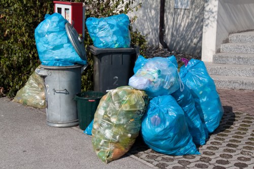 A collection of various overflowing rubbish bags and wheelie bins placed on a paved driveway outside a residential property, with some bags made of blue and green plastic containing mixed waste materials. The bags are piled next to a grey metal bin, a black plastic bin, and a green wheelie bin, all positioned near a garden hedge and a white house wall with concrete stairs. The scene is set in a suburban area, likely within the Chigwell postcode, illustrating typical waste collection and rubbish removal needs. The environment appears bright, with natural daylight illuminating the scene, emphasizing the different textures of plastic bags and the surfaces of the bins. This arrangement reflects common household waste accumulation requiring professional rubbish clearance services such as those offered by House Clearance Chigwell, serving the local community in the vicinity of Essex.