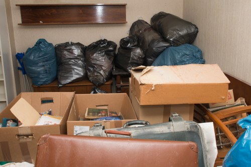 A collection of black and dark blue plastic rubbish bags are stacked on a wooden shelf against a beige wall, with some bags leaning to the sides. In the foreground, several open cardboard boxes contain various discarded items, including papers and small objects, with some boxes partially collapsed or damaged. A large, light brown cardboard box with a torn corner sits atop the boxes, partially covering a grey plastic object which appears to be part of a household item. To the right, a wooden slatted items, possibly a small chair or storage rack, and a blue plastic bag are visible. The scene is indoors, possibly in a garage or storage room on a property in the Chigwell area, indicative of rubbish or household clearance activities conducted by House Clearance Chigwell. The overall environment suggests an organized disposal of waste material awaiting removal or disposal services, with neutral lighting highlighting the various textures and materials present in the cluttered space.