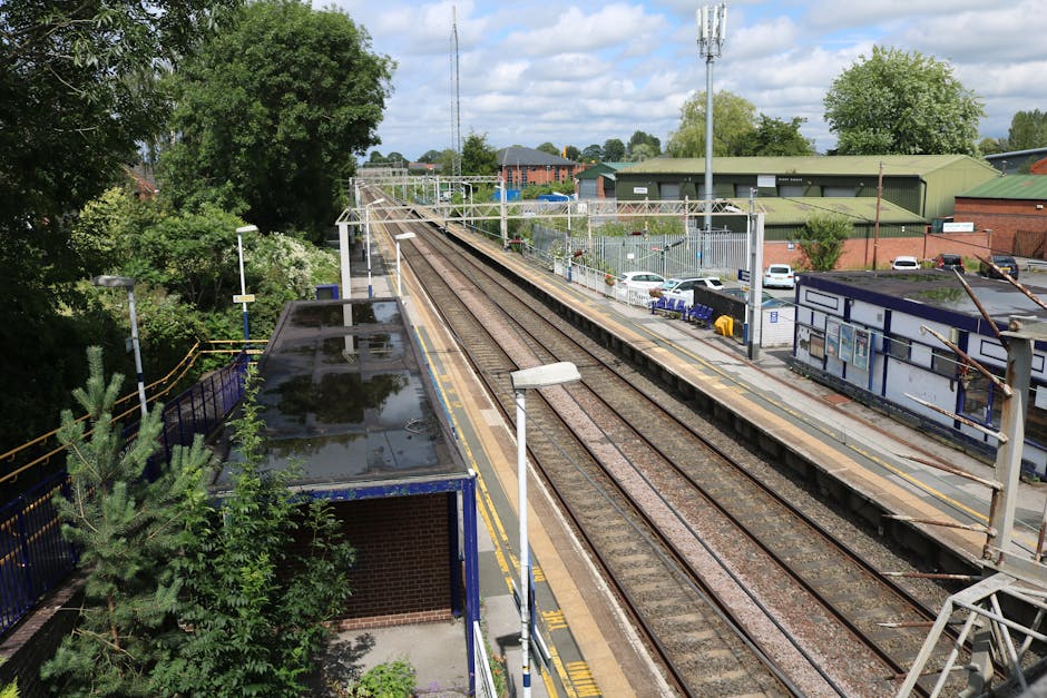 A railway station platform situated in an urban area during daytime, with railway tracks running parallel and bordered by blue safety barriers on the edge. The platform has a shelter structure made of