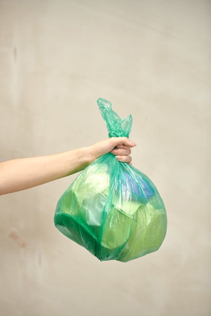 A close-up of a person's hand holding a green plastic rubbish bag by its tied top against a plain, light beige wall. The bag appears partially filled with waste, with some visible folded areas and sli