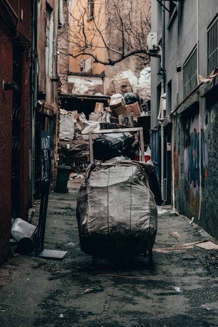 A sanitation worker in a high-visibility orange vest is engaged in waste collection from a bright red rubbish truck parked on the side of a suburban street. The worker is bent over, using a tool to ma